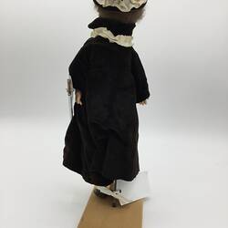 A doll dressed in a cream tunic with a red cross, cream pants and a dark cloak