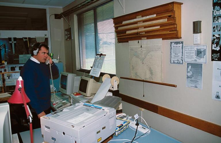 Michael Greenwood wearing remote transceiver headset, at console, with wall-mounted roll-up charts. Melbourne Coastal Radio Station, Cape Schanck, Victoria