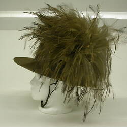 Green khaki cap with plume of feathers on white mannequin head, side view.