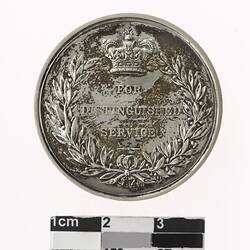 Round silver coloured medal with crown above text and wreath surrounding.