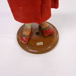 Indian figure attached to a wooden platform in a long red smock wearing flat toed red and green shoes.