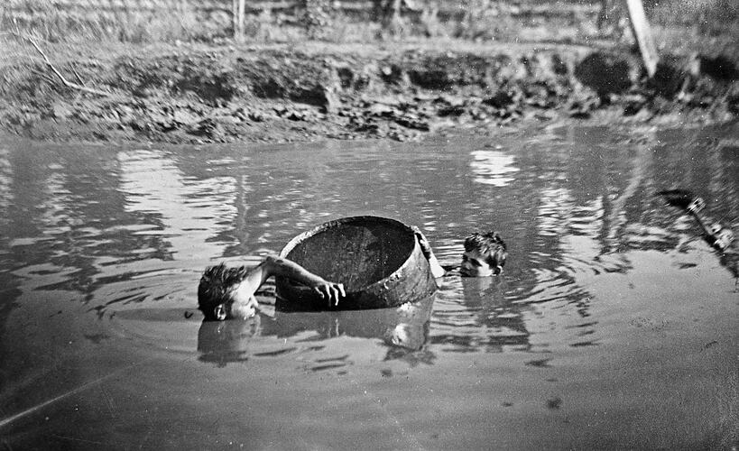 Two boys swimming with a barrel in a water hole.
