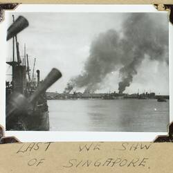 Part of sailing ship on left, land, buildings and billowing smoke in the background.