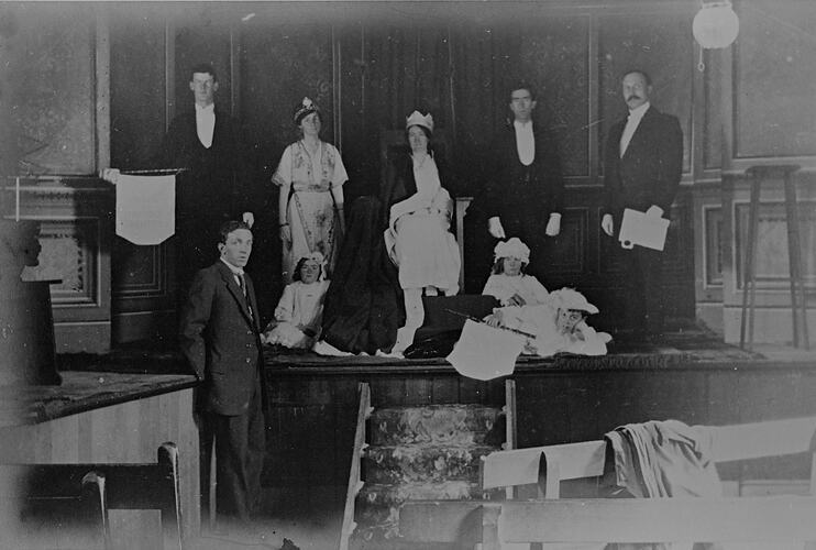 Woman wearing crown seated on stage. Three young children sit either side. Three men and a woman stand either side of her. Man stands on floor below.