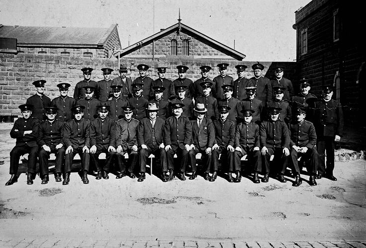 Three rows of male warders in uniform. Bluestone prison wall and jail building behind them.