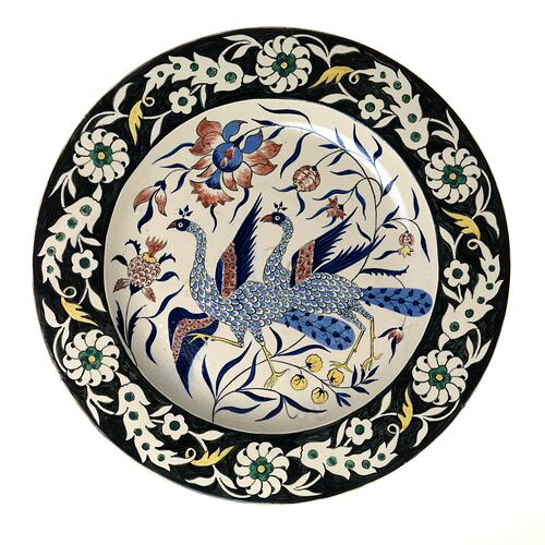 Plate painted with two blue peacocks and flowers in centre. Black rim has white, yellow, green floral motif.