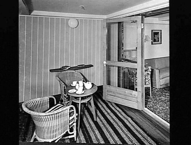 Ship interior. Small semi outdoor area, two round cane chairs around a round table set with a white tea set.