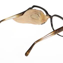 Back view of plastic prosthetic glass left eye behind upper left cheek attached to brown plastic spectacles.