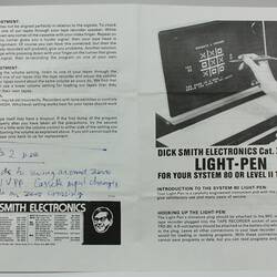 Open white book, black printed text. Image of computer screen (noughts and crosses game) on right. Blue handwriting on left.