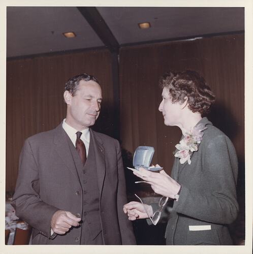 Woman holding glasses talks to man wearing brown.