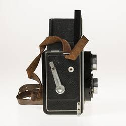 Metal twin lens reflex camera. Body covered in black leather. Leather carry strap. Right profile.