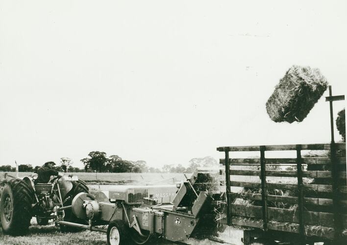 Man driving a tractor with a hay bale thrower with bale in air, loading into a trailer.