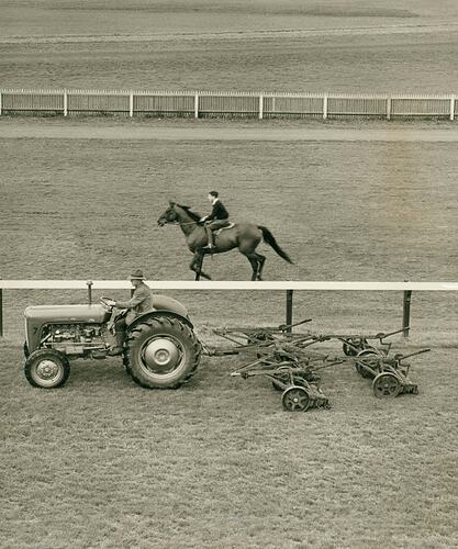 Man driving a tractor at race course, pulling a mower.  Man riding a race horse in background.