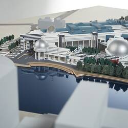 Cardboard model of building with two silver domes and curved steps leading down to river. Viewed across river.
