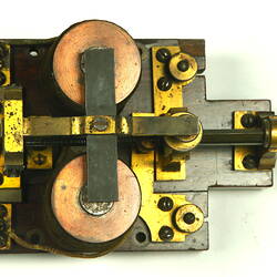 Brass apparatus with battery in centre on wooden base, overhead view.