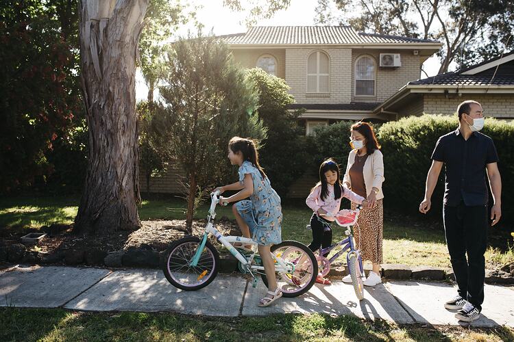 Woman, man and two girls in front of house. The girls are on bicycles. The woman and man wear masks.