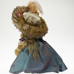 Back of teddy bear wearing lavish cream hat with feather and blue dress jacket.