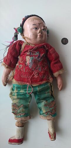 Doll with ceramic head, hands and feet and cloth body. Wears red jacket, green trousers and red shoes.