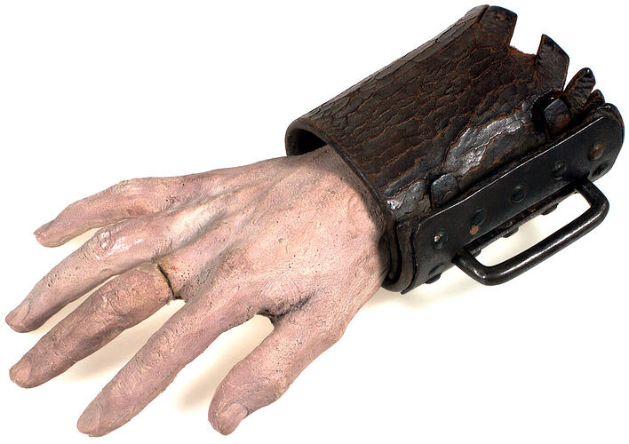 Brown, wide, wrap-around leather wrist shackle on pink painted plaster-cast of male hand and wrist.