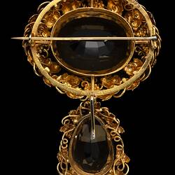 Two part brooch. Top, ornate gold frame with transparent brown cut stone. Below hangs smaller teardrop version
