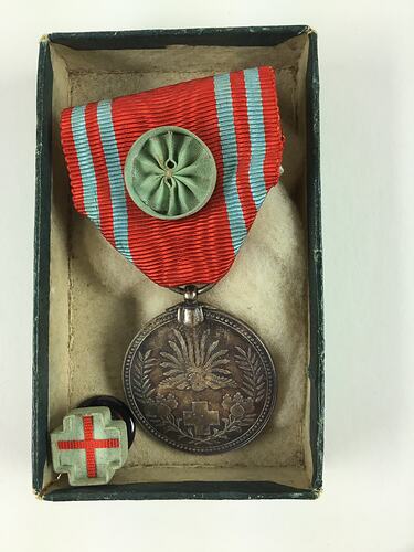 Medal suspended from red ribbon in box. Cross shaped pin with red cross beside it.