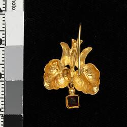Back of gold pendant earring with downward facing bird, two leaves and single red hanging charm.