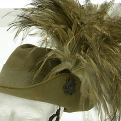 Green khaki cap brim pinned to crown with broach and plume of feathers, on white mannequin head.