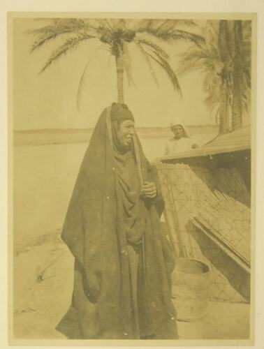 Arab woman wearing chador, palm tree and boy in background.