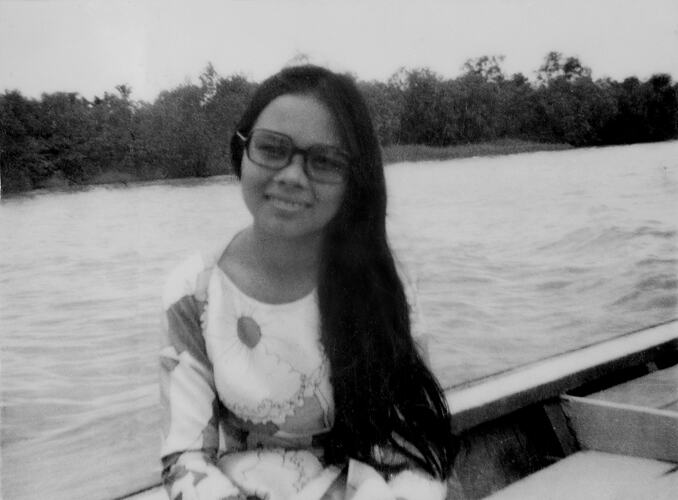 Woman sitting in boat on water, land in background.