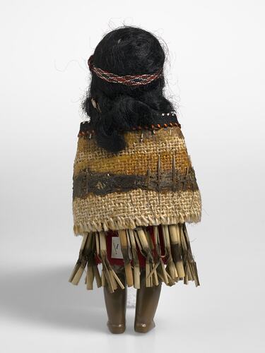 Back of dark brown plastic female doll wearing yellow and brown woven headband, shawl and skirt.