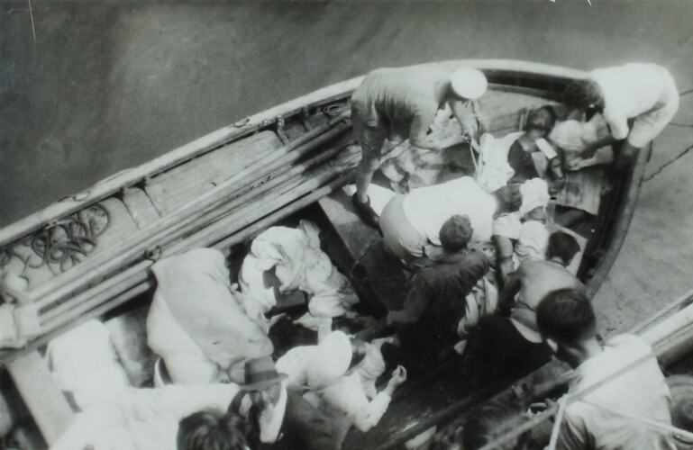 View from above of a lifeboat with multiple people assisting a wounded man laying at bow of boat.