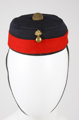 Dark blue pillbox hat with red band around edge. Gold insignia on front, gold braid button on crown, chin stra
