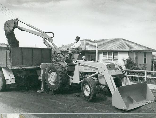 Man operating a backhoe loader, filling up a truck with soil, in front of a house,