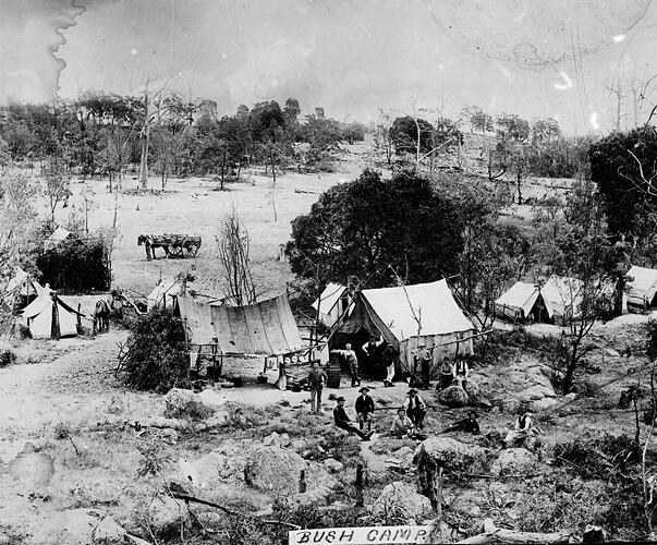 Tents set up beside a creek. Men in foreground, horse and dray in clearing behind them.