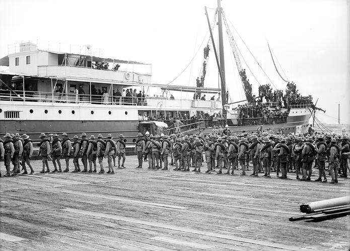 Crowd of soldiers wharf with ship with crowded bow behind.