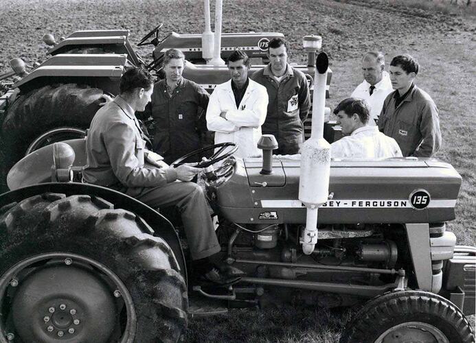Group of men stand between tractors in field.