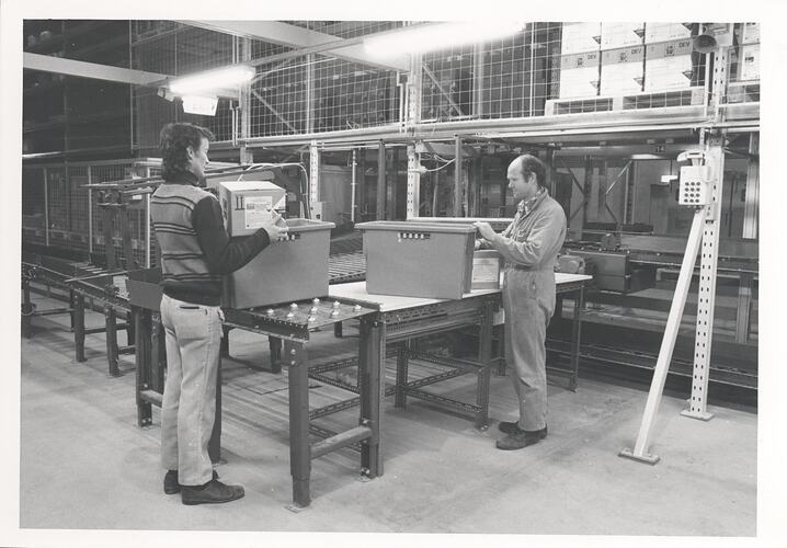 Two men with boxes near warehouse conveyor belt.