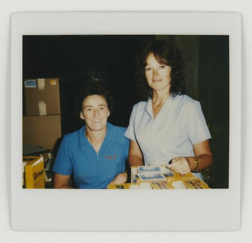 Two women pose, one is sitting at left and the other is standing at right. They are surrounded by boxes.