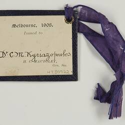 Dark blue card with white label bearing printed and handwritten text. Attached to a frayed purple ribbon.