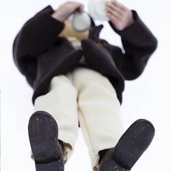 Miniature man in dark coat, light pants. Holds a cup and saucer. Has a hat and fair hair. View from below.