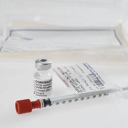 Labelled syringe with vial behind it and plastic box in background.