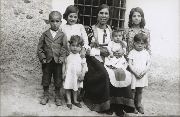 Seated woman poses with six young children. Dirt wall and metal grid window behind them.