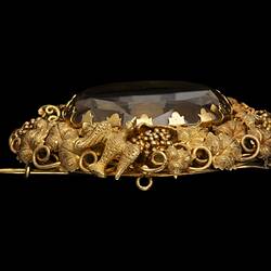 Profile of two part brooch. Top, ornate gold frame with transparent brown cut stone. Below hangs smaller teardrop version