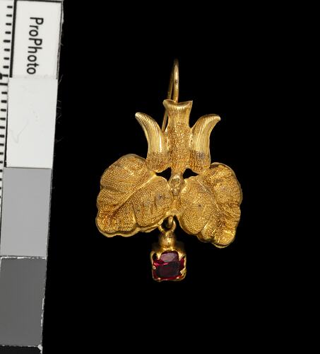 Gold pendant earring with downward facing bird, two leaves and single red hanging charm.