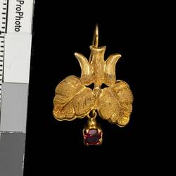 Gold pendant earring with downward facing bird, two leaves and single red hanging charm.
