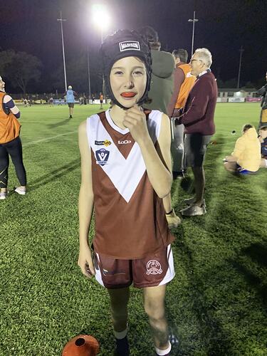 Half portrait, girl wearing maroon and white V design football jumper and blue helmet. Night time on oval with lights on.