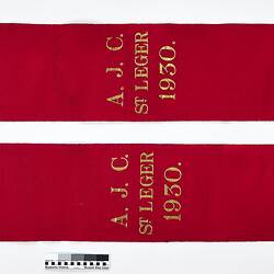 Folded red ribbon sash with gold lettering and fringe.
