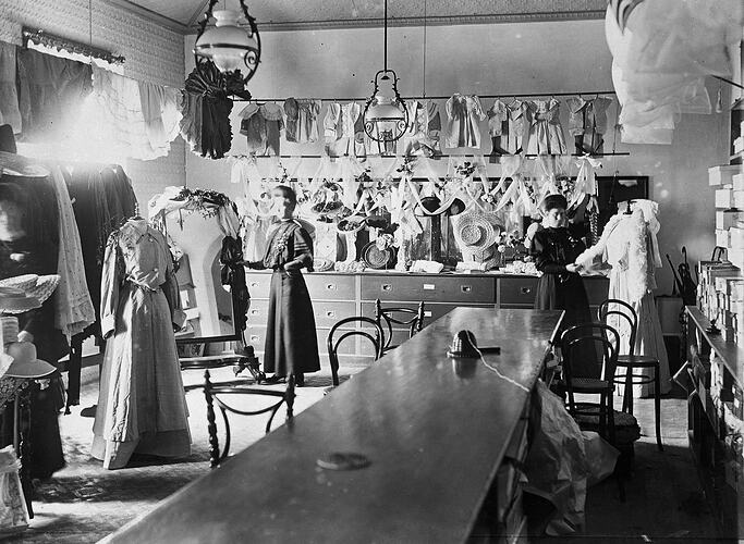 Ladies clothing shop. Central long wooden bench.  Victorian era dresses on mannequins. Two women in similar at
