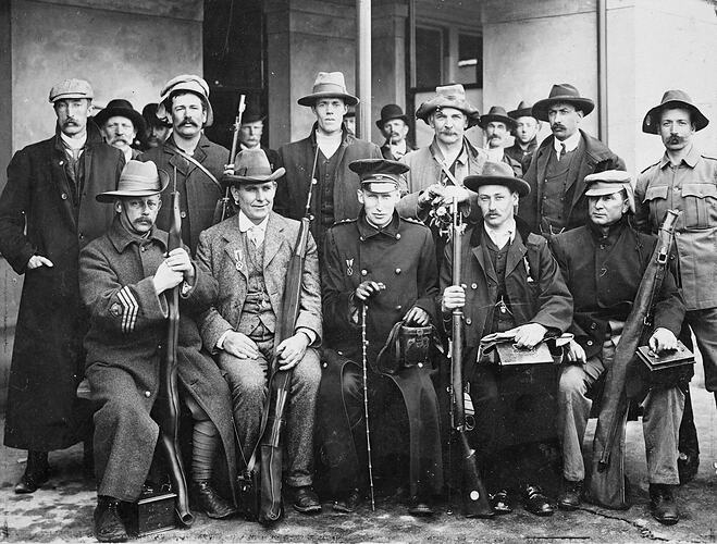 Row of seated men holding rifles with row standing behind them. In front of building.