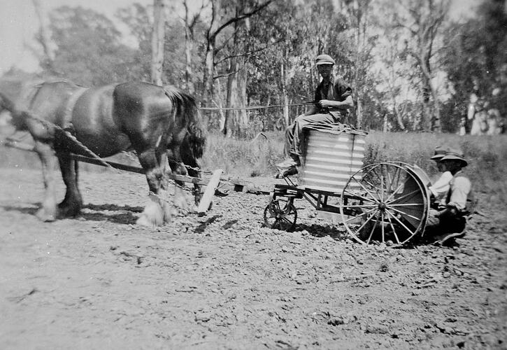 Two horse drawn cart in field with driver sitting on metal corrugated tank. Two men are seated behind the cart close to the ground.